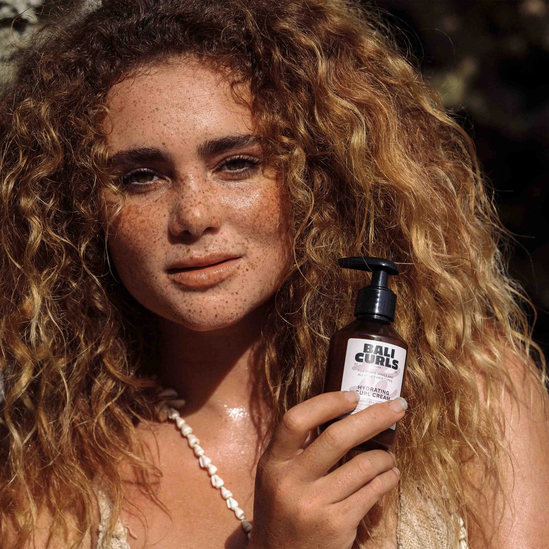 Een persoon met krullend haar houdt een flesje Bali Curls Hydrating Curl Cream 150ml (FULL-SIZE) met sheaboter vast.