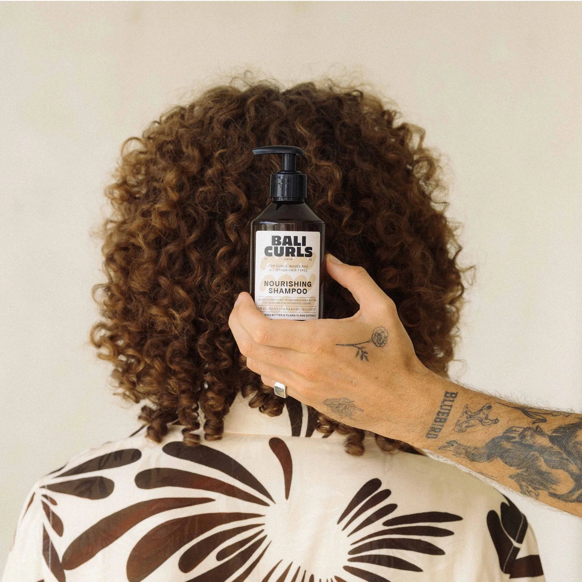 Een persoon met krullend haar en een shirt met een printje houdt een fles van 250 ml Bali Curls Nourishing Shampoo met sheaboter vast.