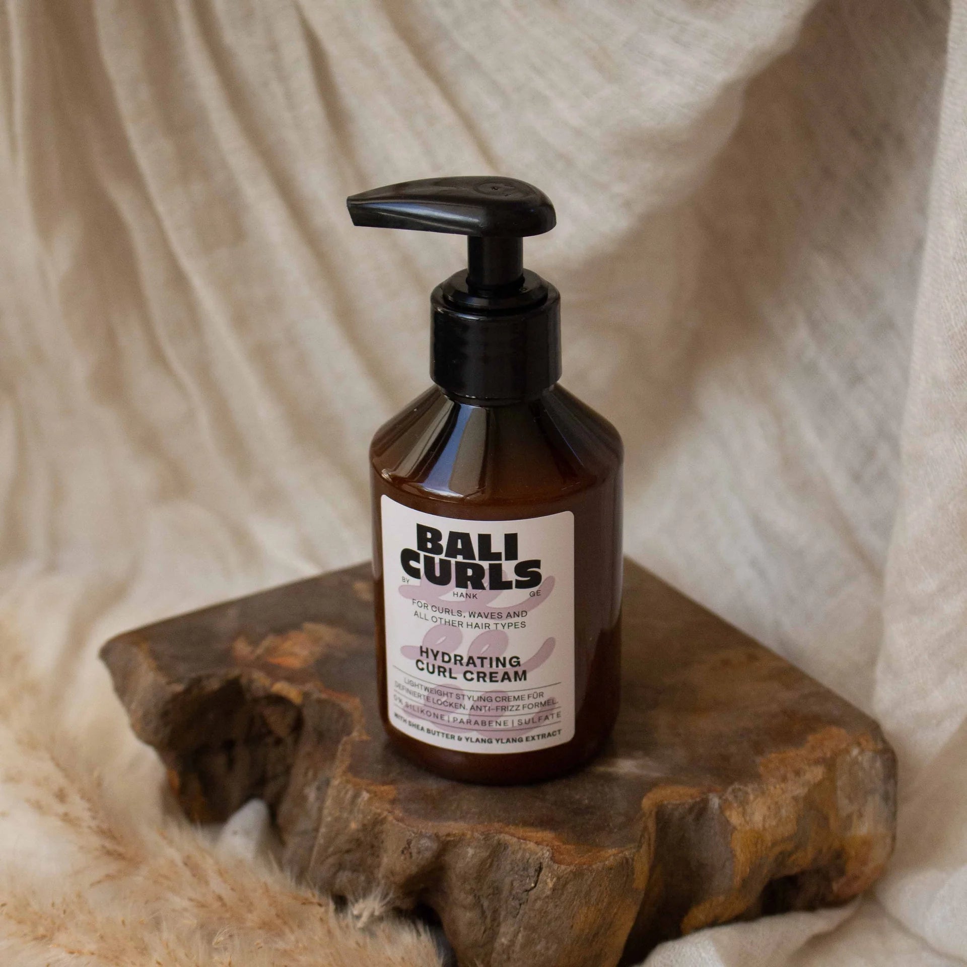 Een flesje van 150 ml Bali Curls Hydrating Curl Cream, compleet met pompje en verrijkt met sheaboter voor extra voeding, staat op een houten oppervlak tegen een beige stoffen achtergrond.
