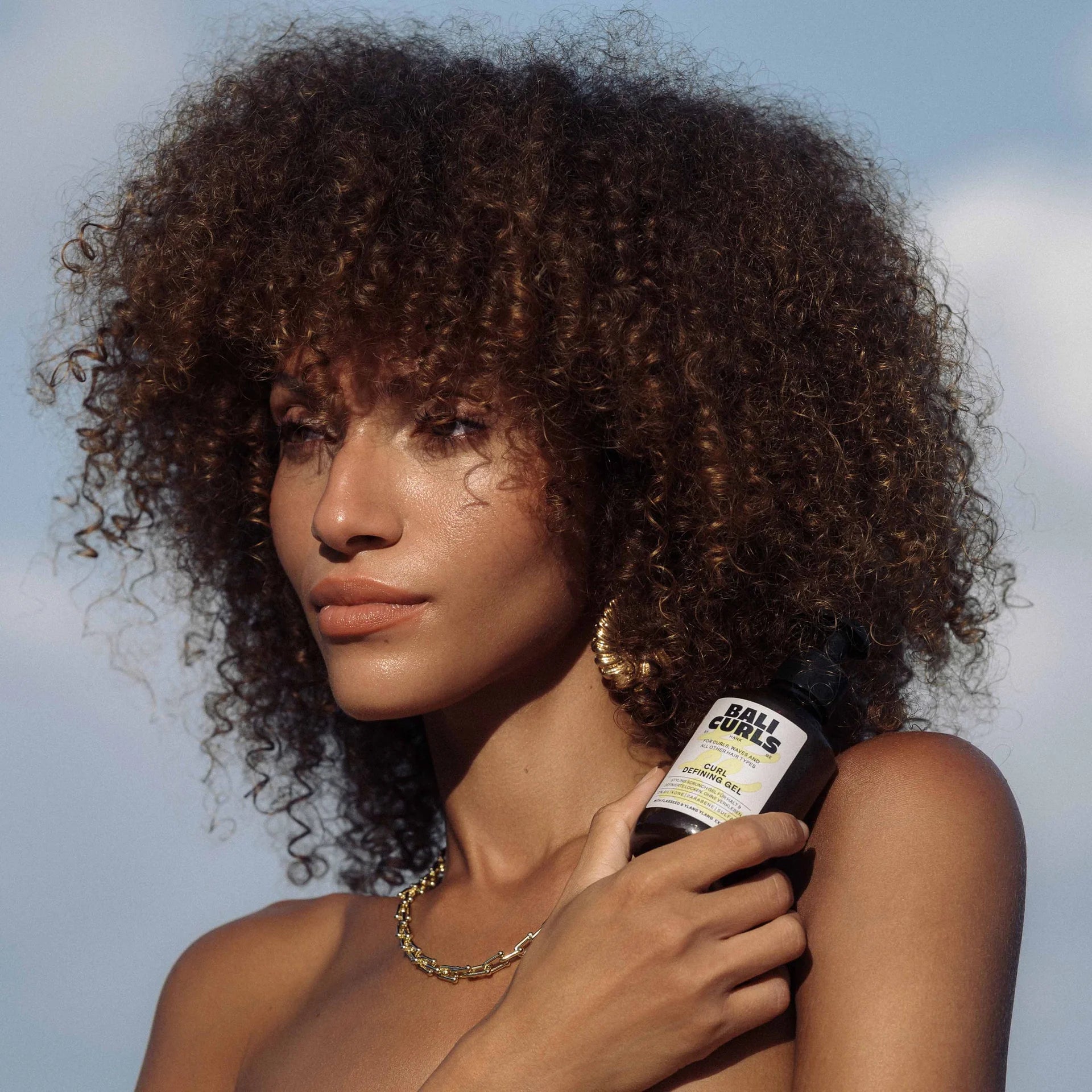Een persoon met krullend haar houdt een fles vast met het label Bali Curls Curl Defining Gel 150ml (FULL-SIZE). Ze dragen een gouden ketting en oorringen en staan tegen een lichtblauwe lucht met wolken.