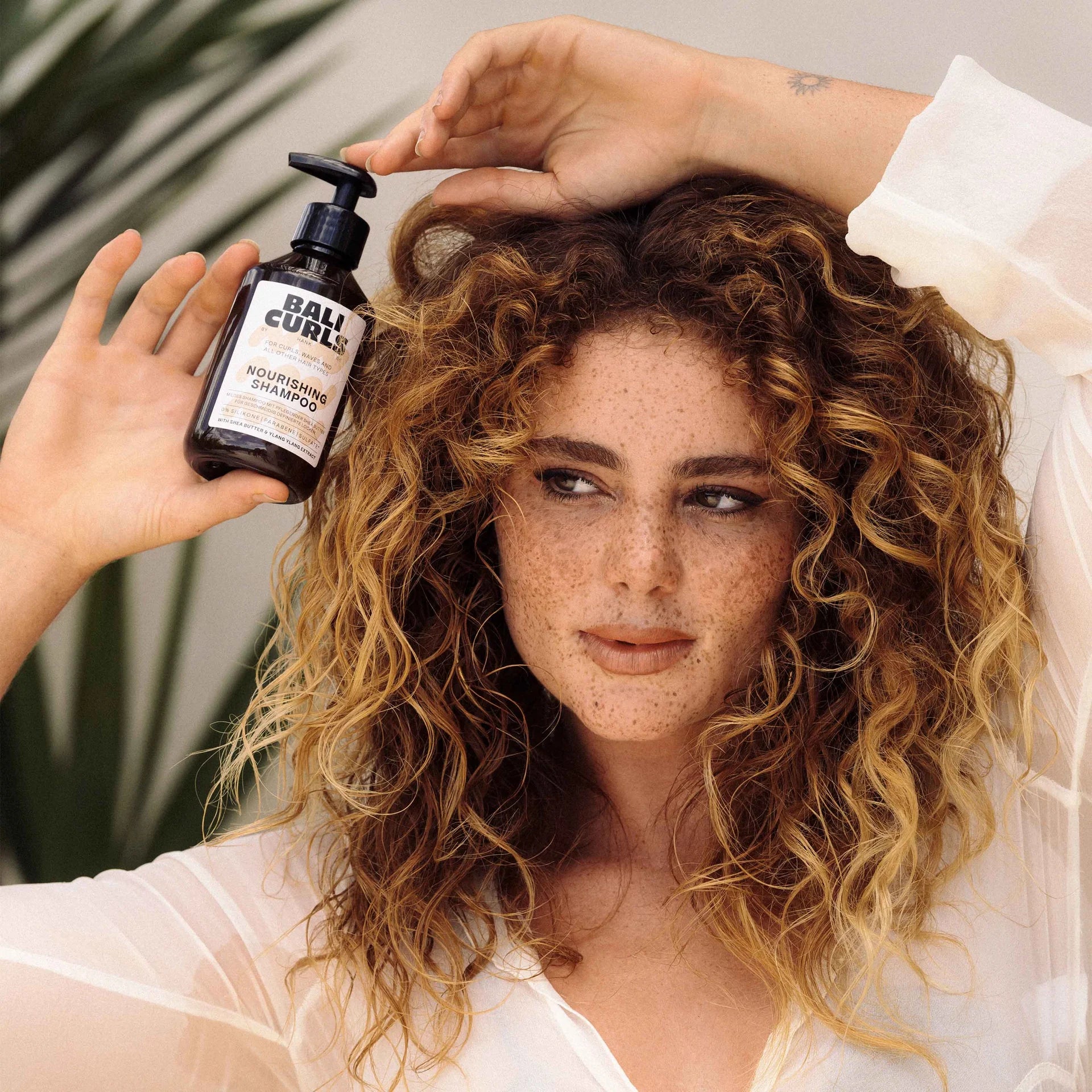 Iemand met krullend haar houdt een fles Bali Curls Nourishing Shampoo 250ml (FULL-SIZE) vast. Deze is verrijkt met sheaboter voor ultieme hydratatie.