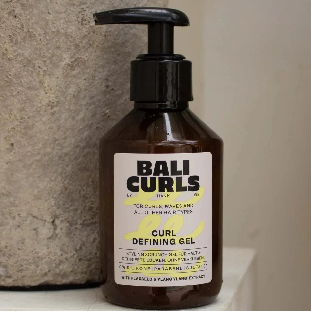 Een fles Bali Curls Curl Defining Gel 150ml (FULL-SIZE) met een handige pompdispenser staat elegant tussen twee stenen vazen. Het etiket toont de veelzijdigheid voor krullen, golven en andere haartypes, verrijkt met lijnzaadextract, chia-extract en crème deluxe.